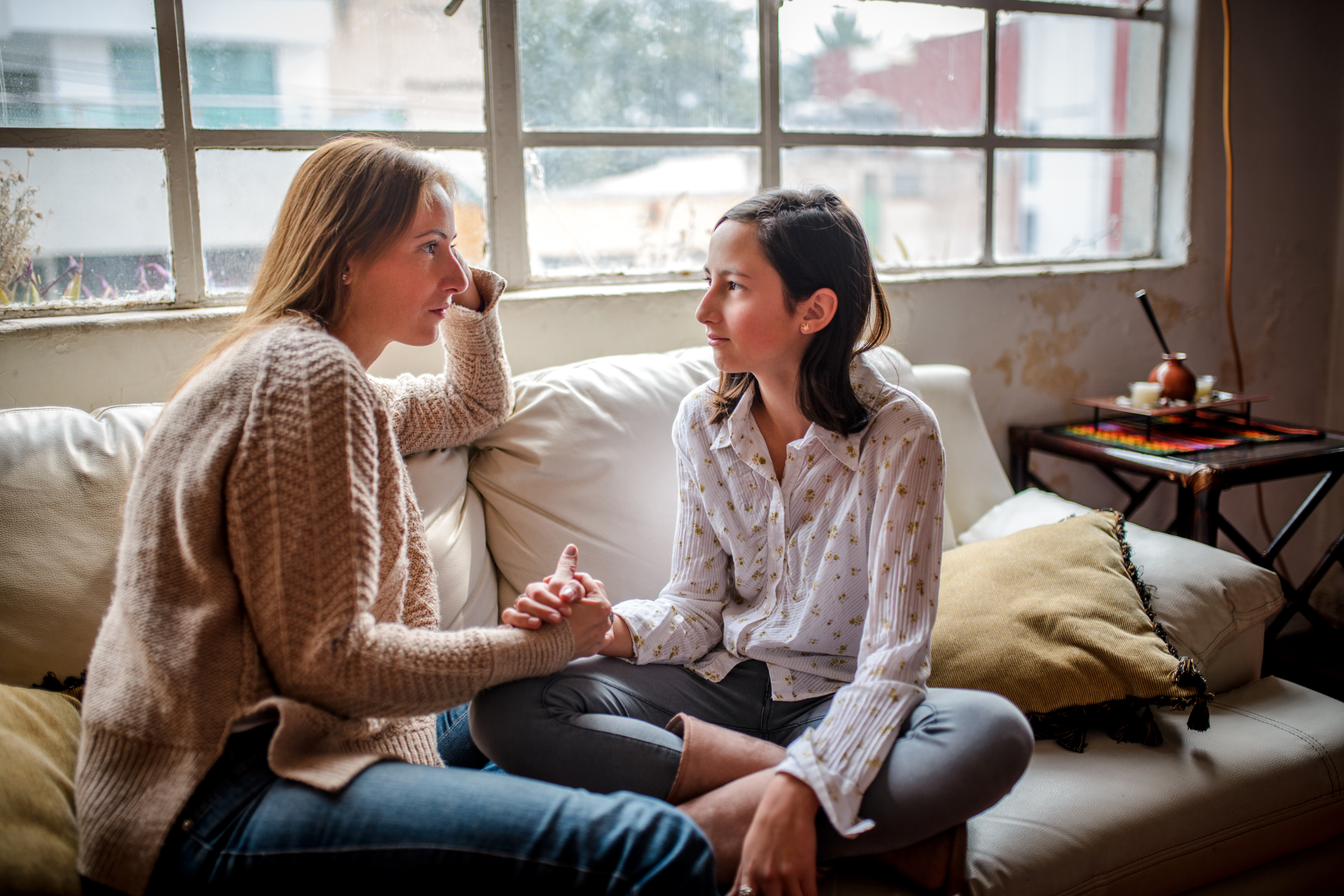 a mother and a teen daughter sit calmly on a couch holding hands